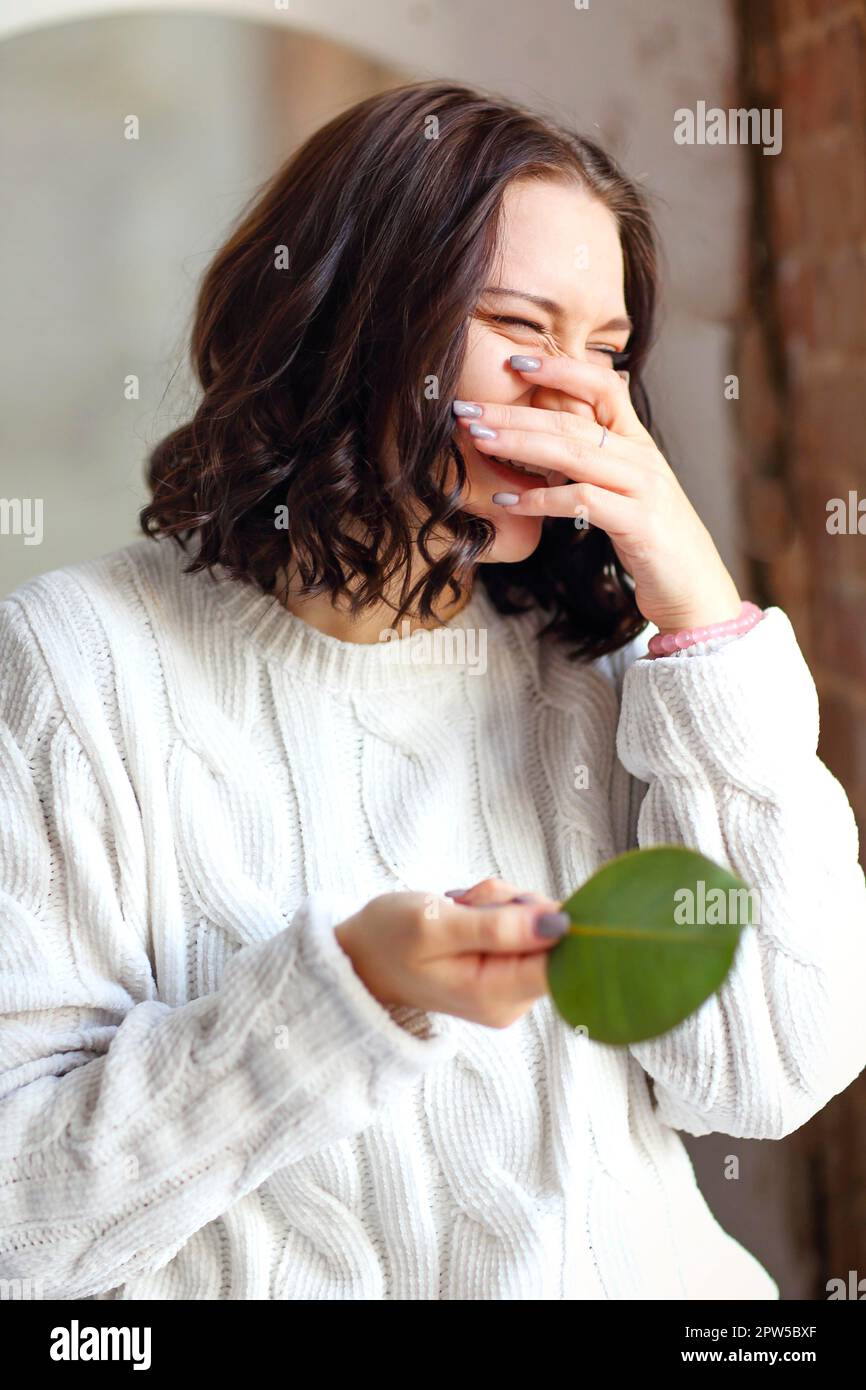 Ritratto di giovane ragazza brunette con capelli splendidamente styled in bianco caldo maglione a maglia, tenendo verde foglia di pianta, in posa contro appartamento interno Foto Stock