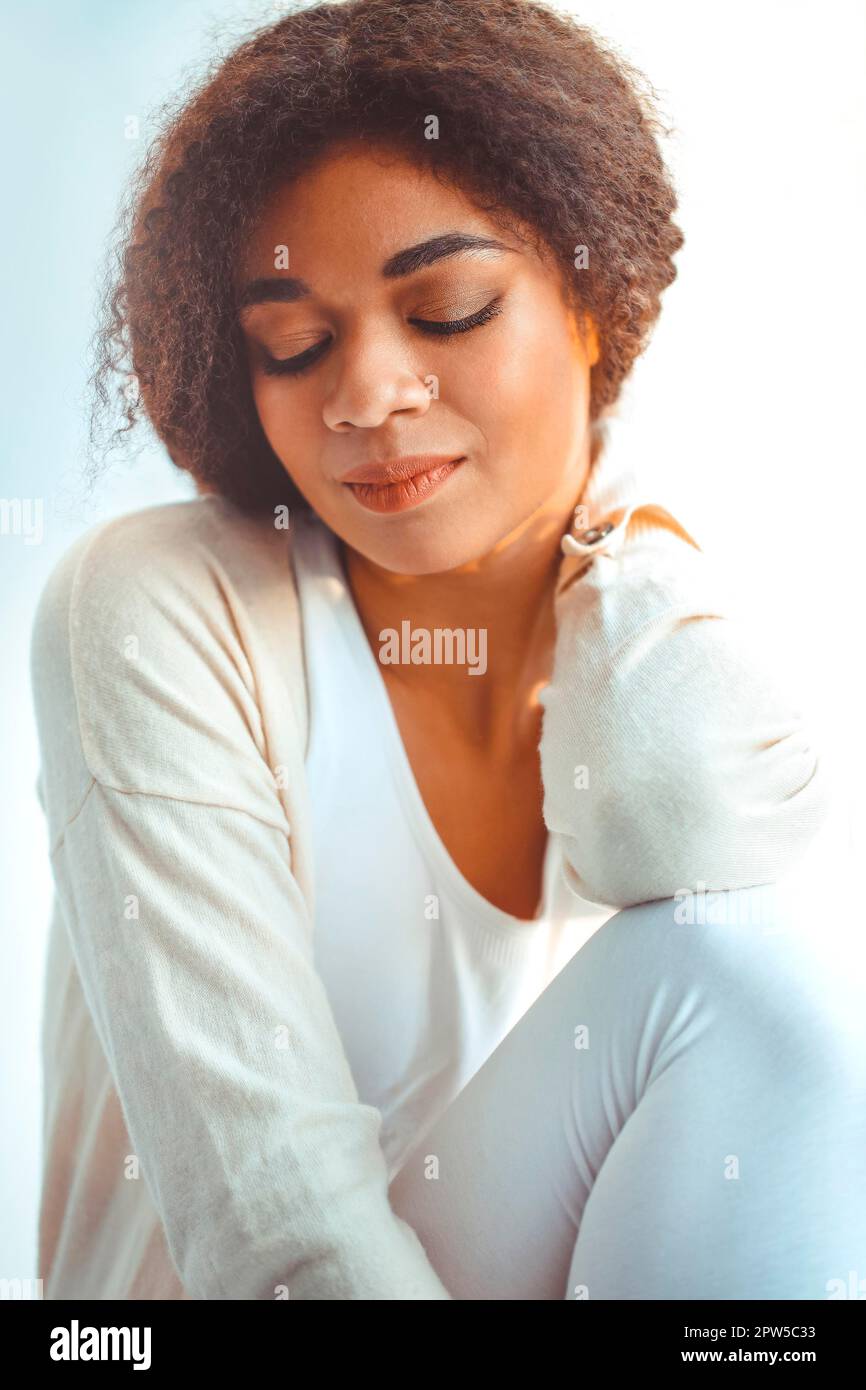 Felice African American ragazza etnia rilassato in abiti da casa seduti su davanzale sullo sfondo di tende che ondolano dal vento chiaro, felice afro Foto Stock