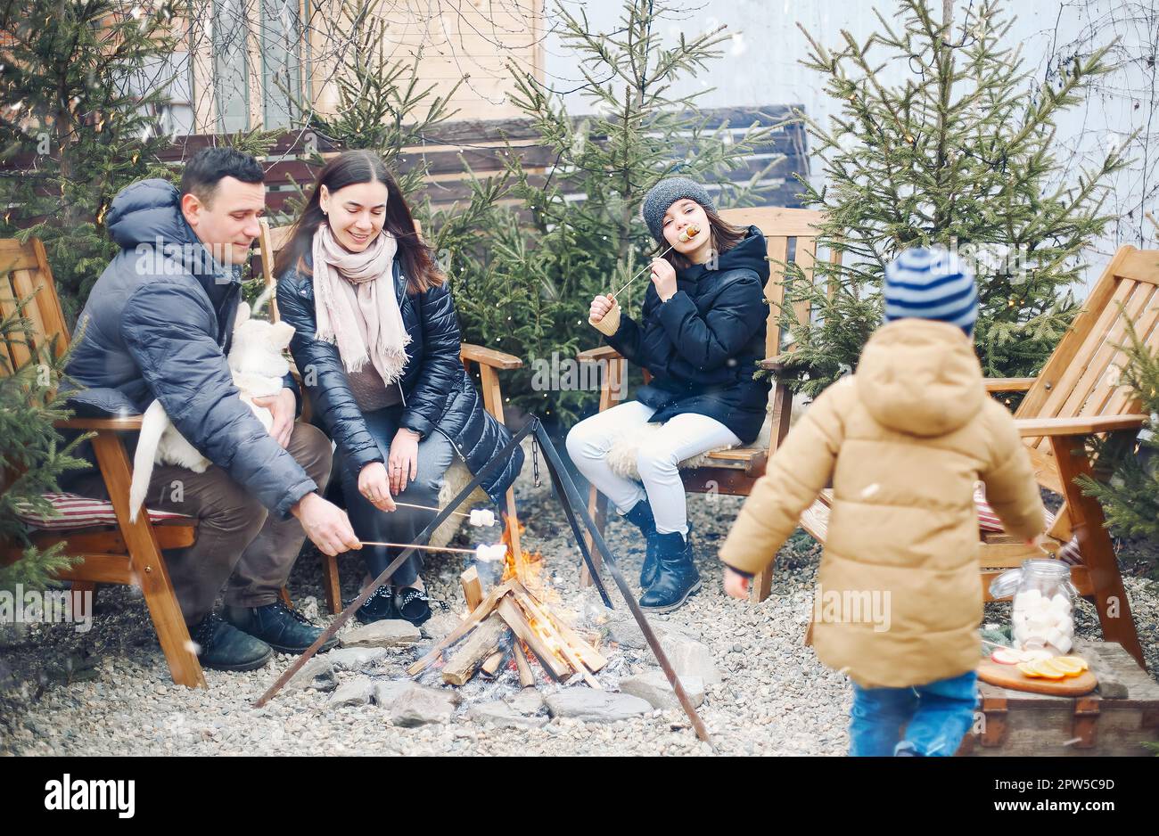 Famiglia in abiti caldi seduti attorno al fuoco del campo acceso all'esterno, arrostire marshmallows su bastoni e chiacchierare, foto ritagliata. Passatempo piacevole Foto Stock