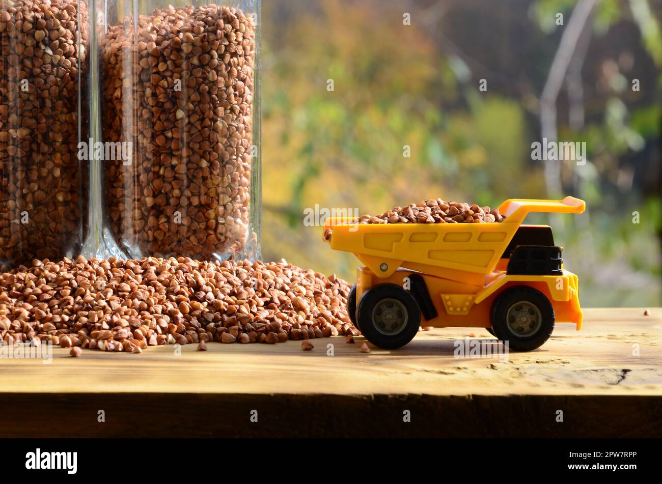 Un piccolo giocattolo giallo carrello è caricato marrone con i chicchi di grano saraceno intorno al mucchio di grano saraceno e un bicchiere della groppa. Una macchina su di una superficie di legno contro un Foto Stock