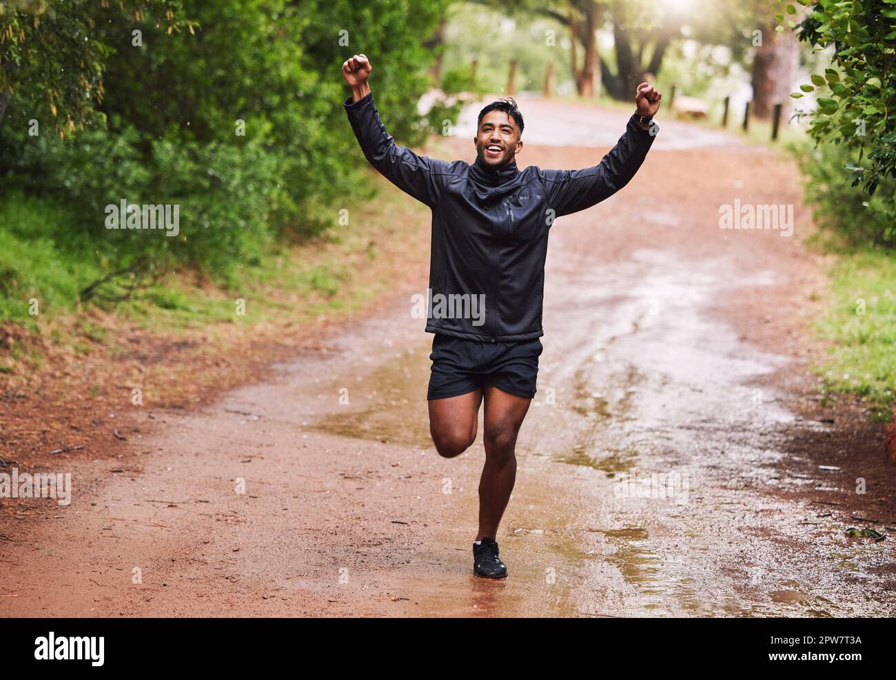 Giovane atleta ispanico vestito maschile che allieta con i suoi pugni in aria, mentre in una corsa in una foresta sotto la pioggia fuori in natura. L'esercitazione è buona per guarire Foto Stock