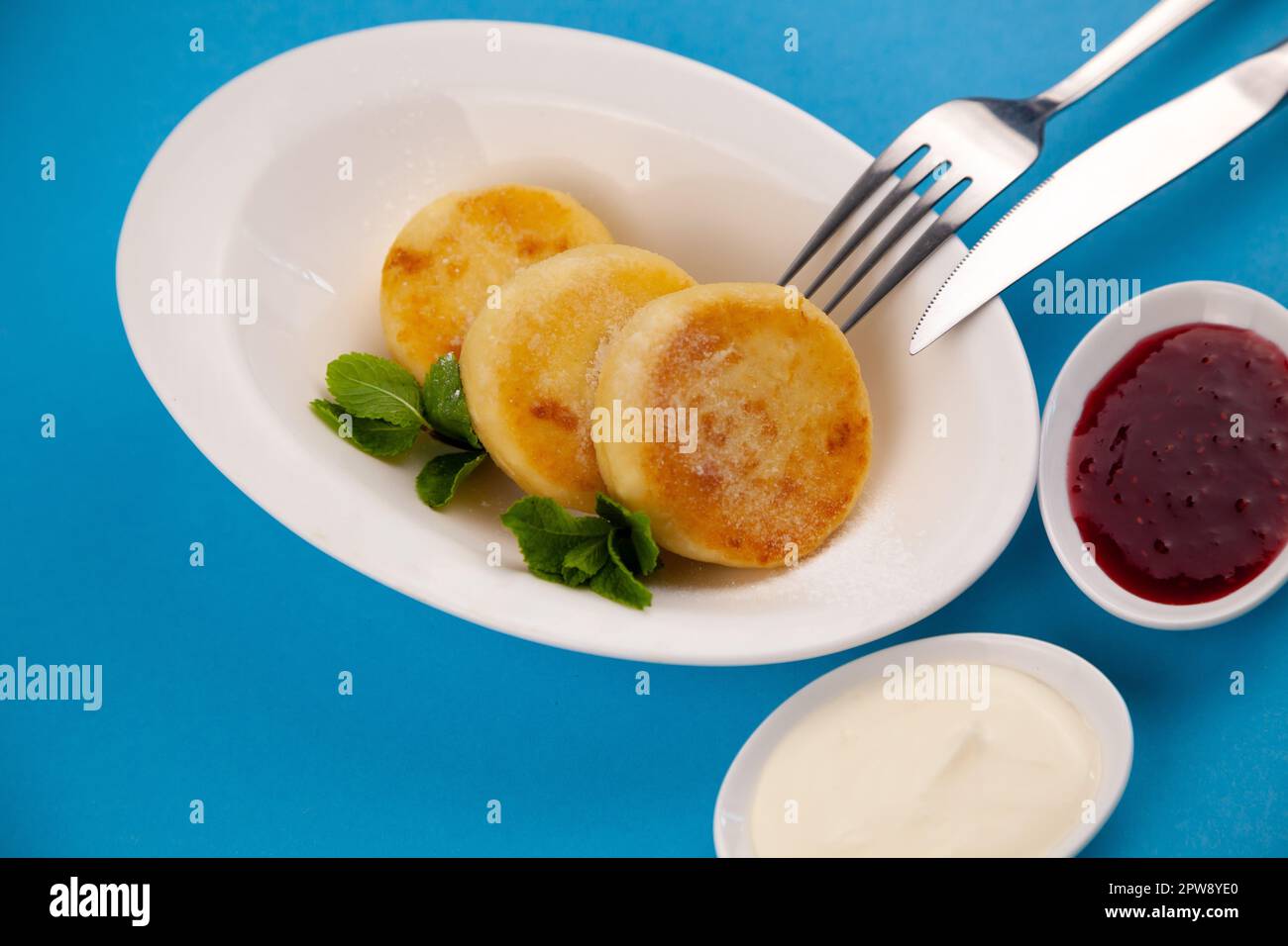 Frittelle di formaggio ricotta rotonda in un piatto bianco su sfondo blu. Cibo prima colazione. Cibo sano. Salsa di panna acida e menta. Primo piano vista dall'alto. Pasto dessert. Frittelle di ricotta al formaggio ricotta. Foto Stock