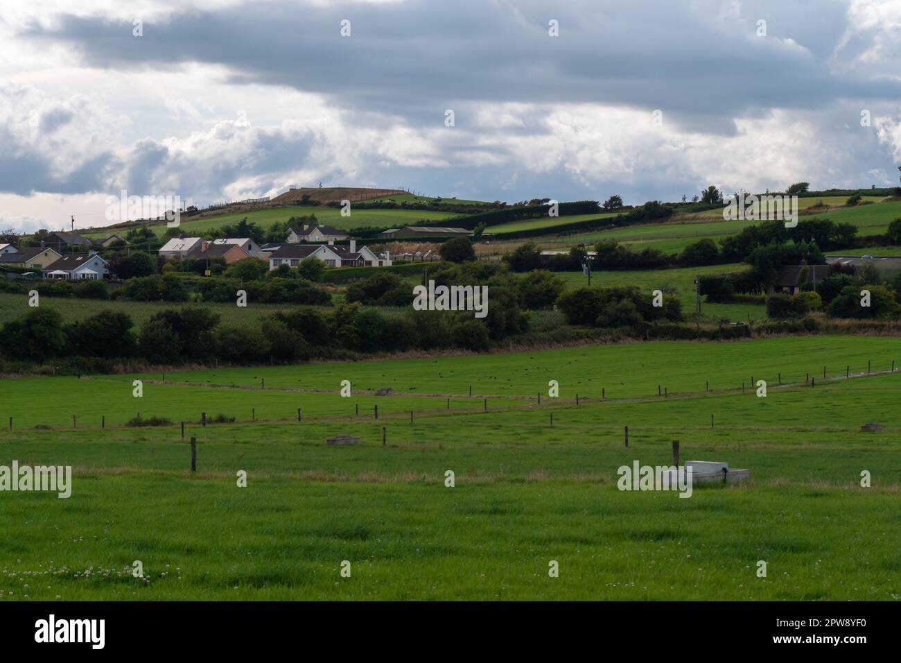 Un piccolo villaggio tra campi e alberi sotto un cielo nuvoloso sera, County Cork. Rustico paesaggio irlandese in estate, villaggio europeo. Prato verde Foto Stock