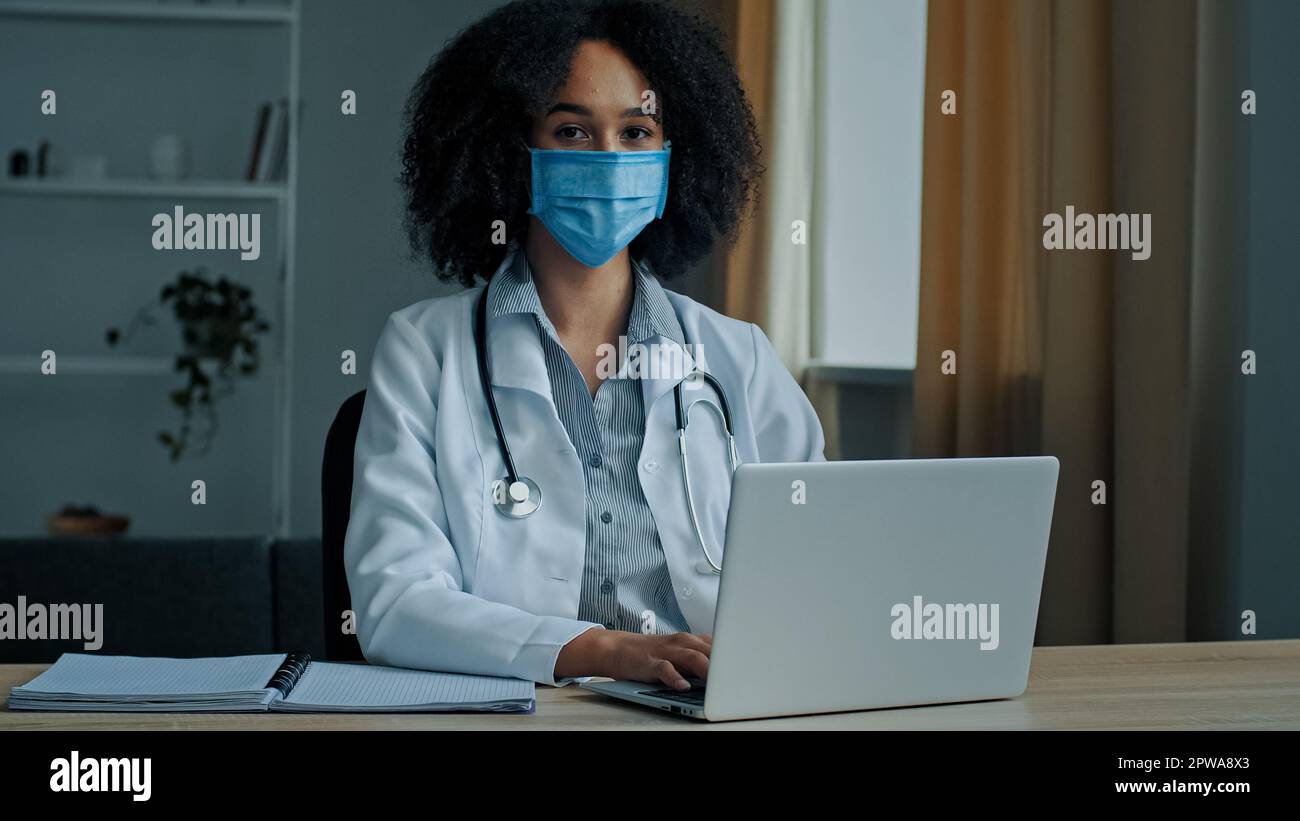 Etnia africana donna medico in maschera viso uniforme sedersi a ospedale armadio lavoro sul computer portatile fare ricerca medica consultazione controllo elettronico a distanza Foto Stock