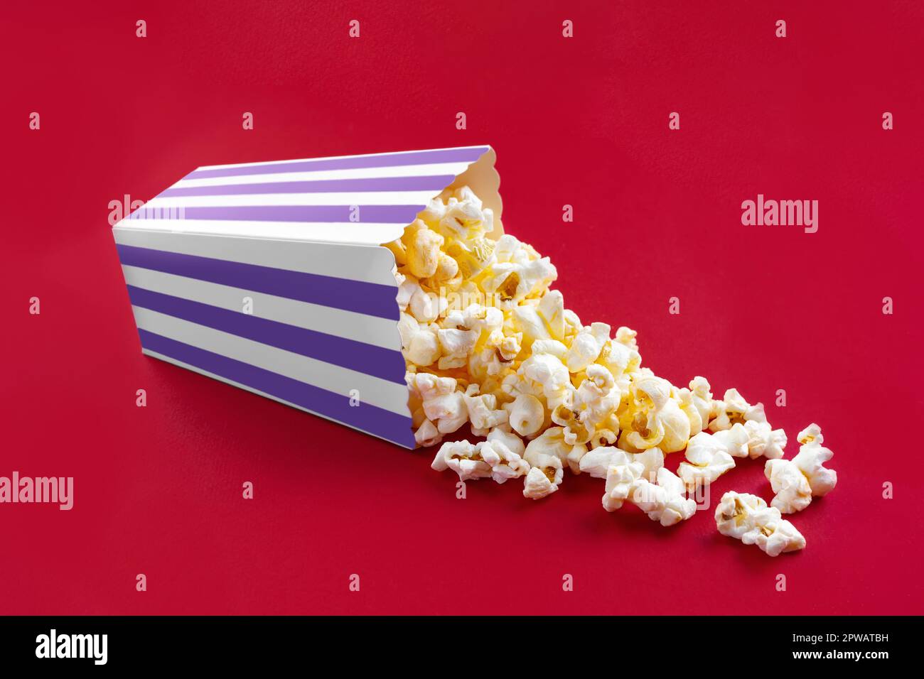 Gustoso popcorn al formaggio che cade da un secchio di cartone a righe viola, isolato su sfondo rosso. Dispersione di grani di popcorn. Film, cinema e ingresso Foto Stock