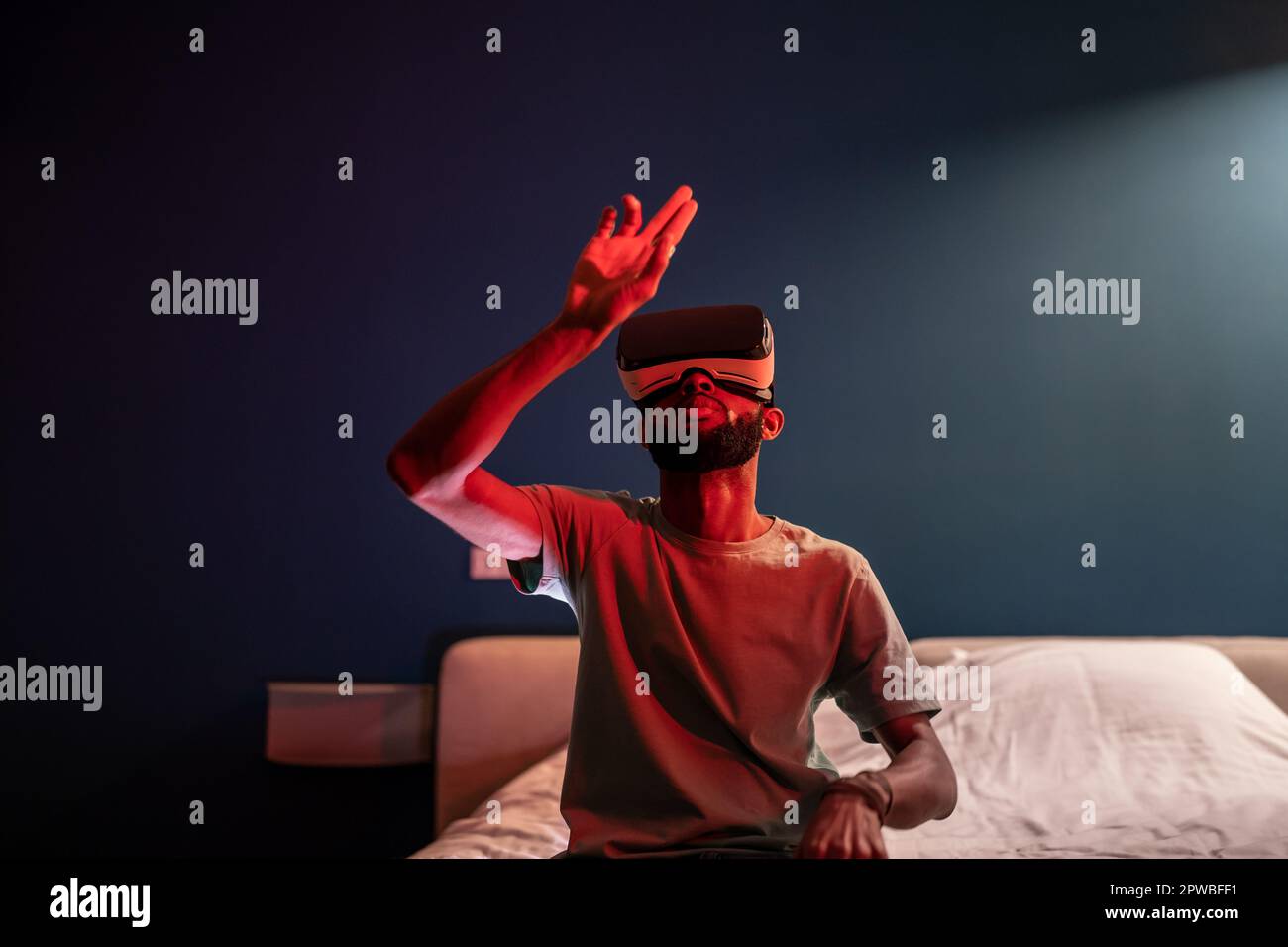 Giovane afro-americano seduto sul letto sotto la luce rossa al neon utilizzando futuristici metaverse VR visore Foto Stock