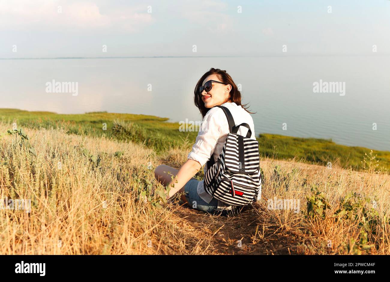 Vista laterale del viaggiatore femminile di brunette seduto sull'erba hill e ammirare paesaggi incredibili durante il tramonto in estate Foto Stock