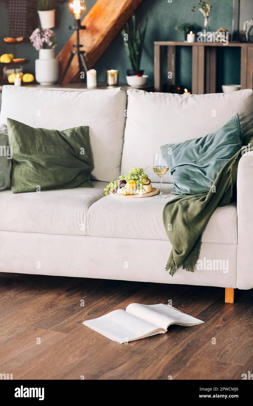 Piatto con frutta fresca e bicchiere di vino collocato vicino ai cuscini su un comodo divano nell'elegante camera di casa Foto Stock