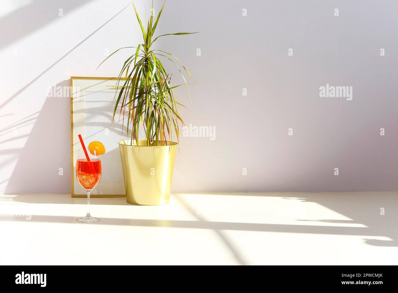Bicchiere di cocktail di frutta fresca collocato al piano vicino alla pianta in vaso e cornice vuota contro la parete bianca nella stanza illuminata dal sole Foto Stock