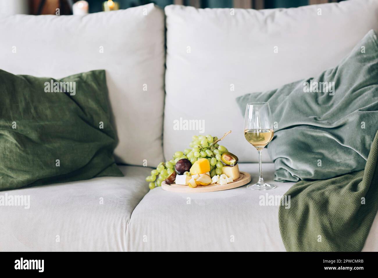 Piatto con frutta fresca e bicchiere di vino collocato vicino ai cuscini su un comodo divano nell'elegante camera di casa Foto Stock