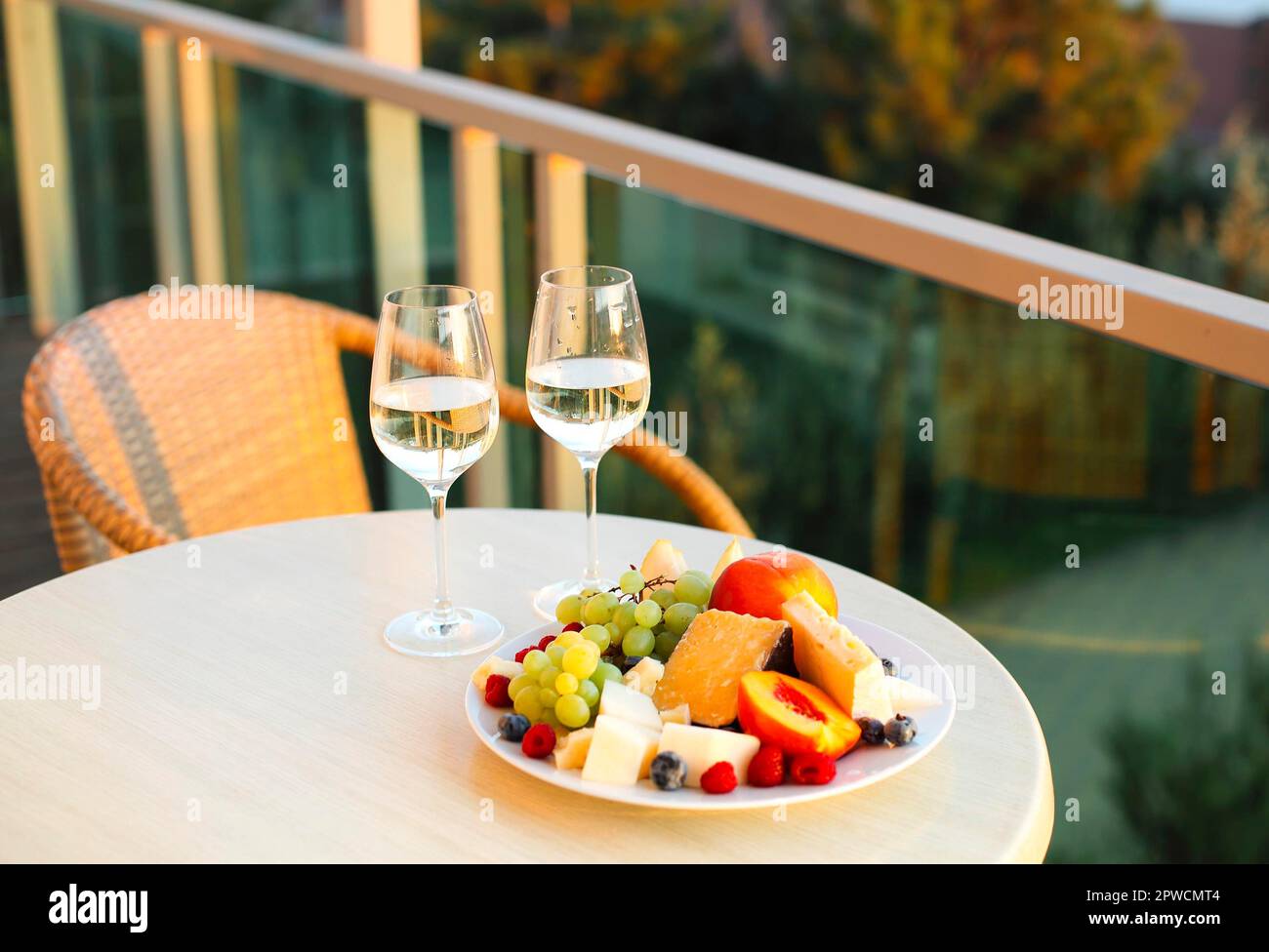 Taglie di formaggio assortite e un piatto di frutta con due bicchieri di vino bianco sul tavolo rotondo che si erge sul balcone dell'hotel affacciato sulla foresta estiva Foto Stock