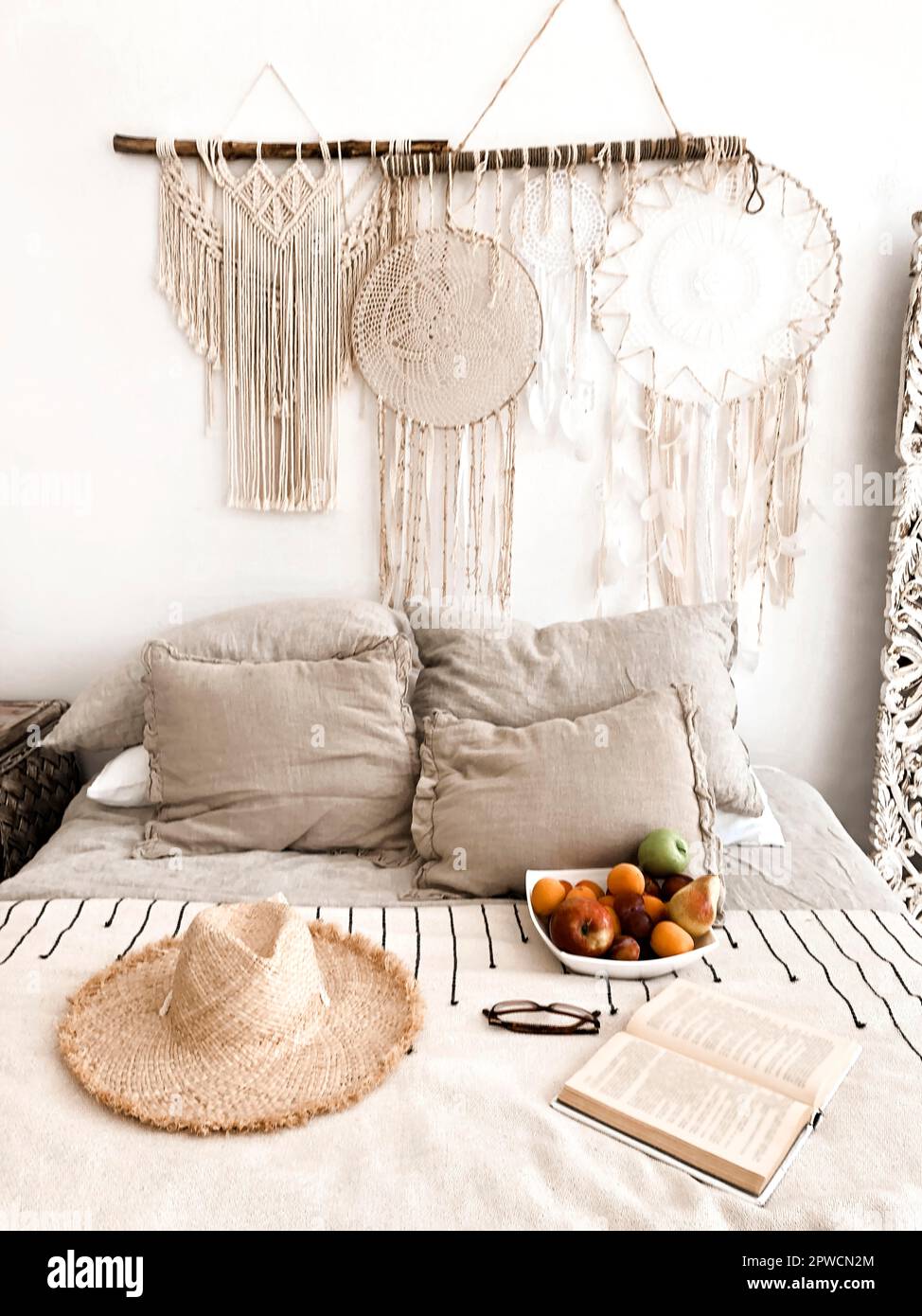 Interni moderni in stile scandi boho con cuscini decorativi, cappello in vimini, frutta e libro aperto sul letto, decorazioni in macrame e dreamcatcher Foto Stock