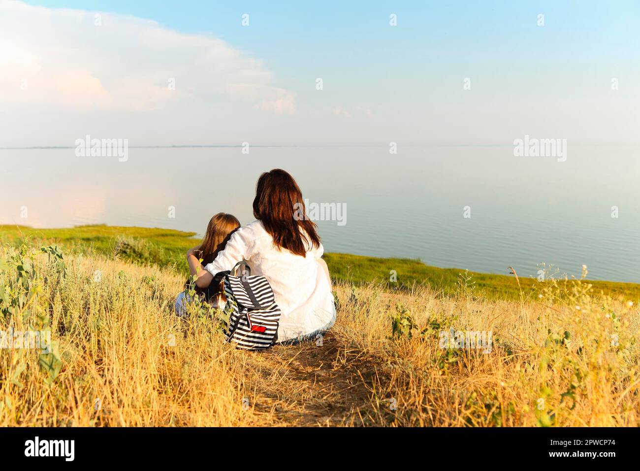 Vista posteriore di donna irriconoscibile abbracciando la ragazza mentre si siede sopra erba secca e ammirando il pittoresco paesaggio marino durante il fine settimana estivo Foto Stock