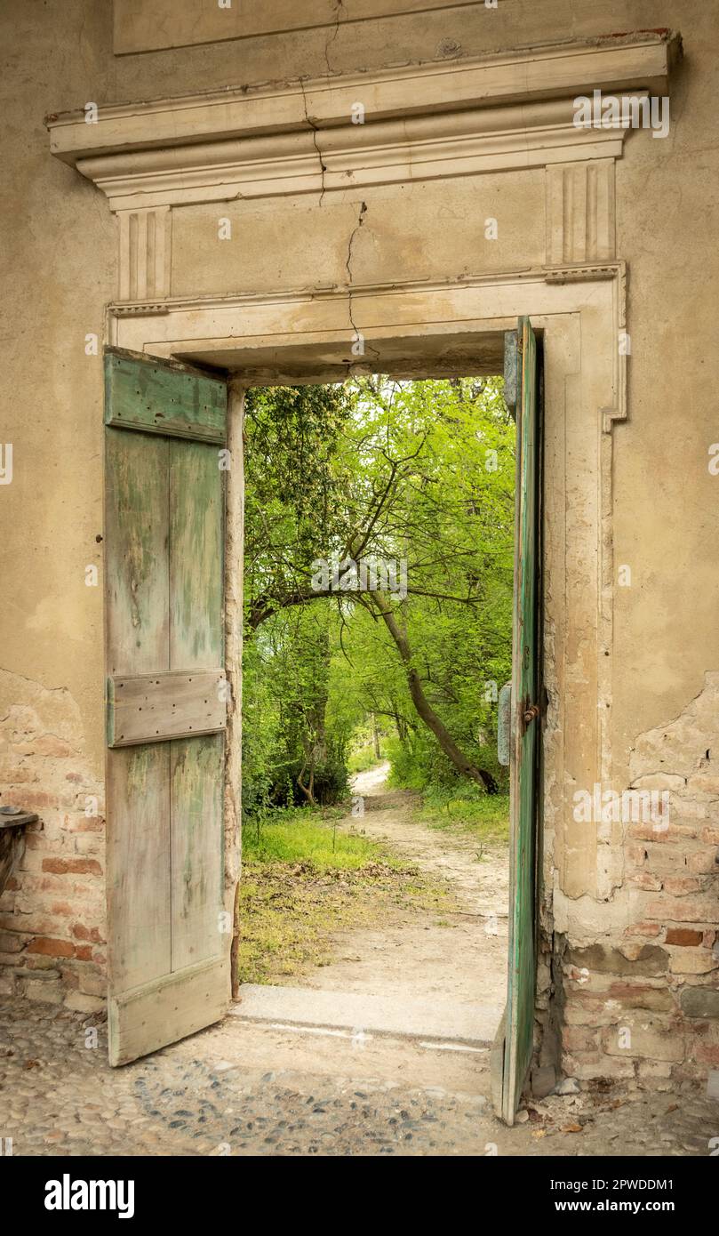 Splendida vista su un parco o boschi con vegetazione lussureggiante attraverso una porta aperta in legno e un muro di pietra di un antico castello di San Sebastiano da po Foto Stock