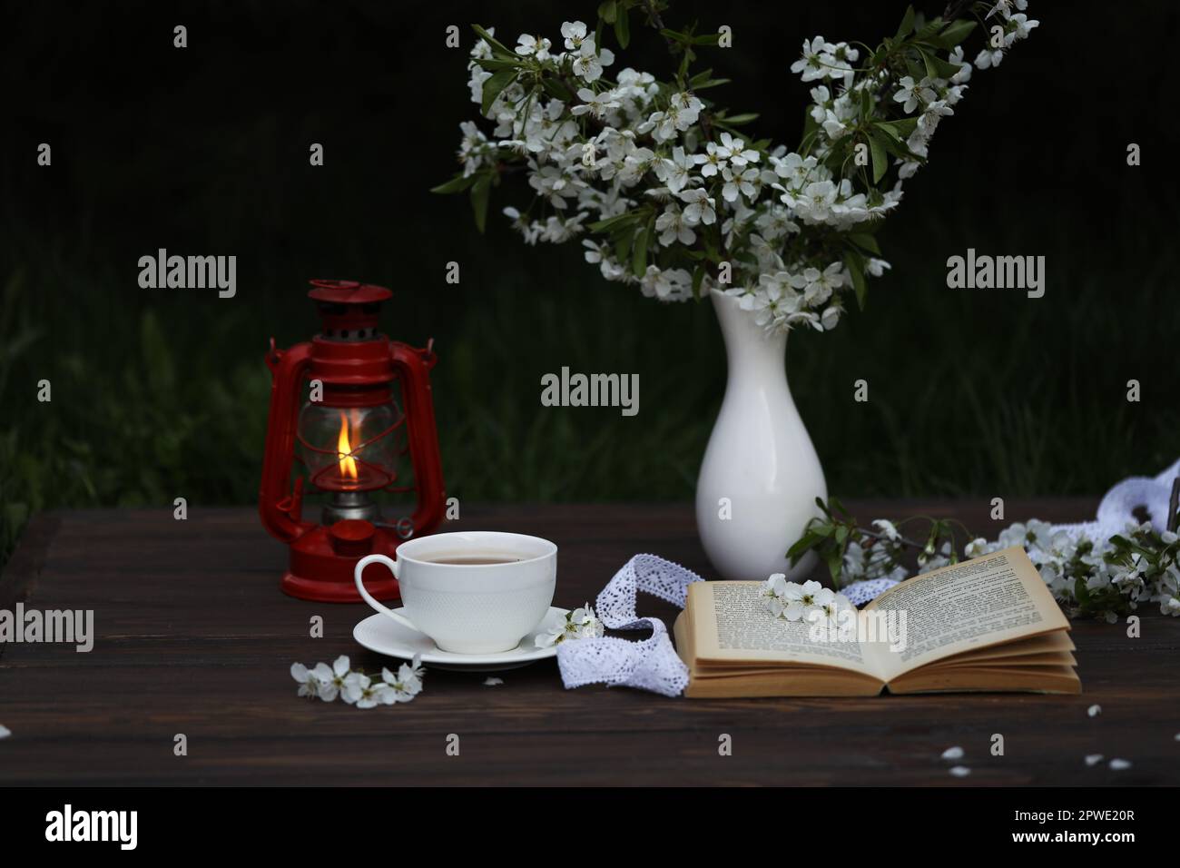 tazza di tè con decorazioni in fiore di ciliegio su sfondo di legno marrone scuro. Chokolate, bouquet, libro e lampada kerosene Foto Stock