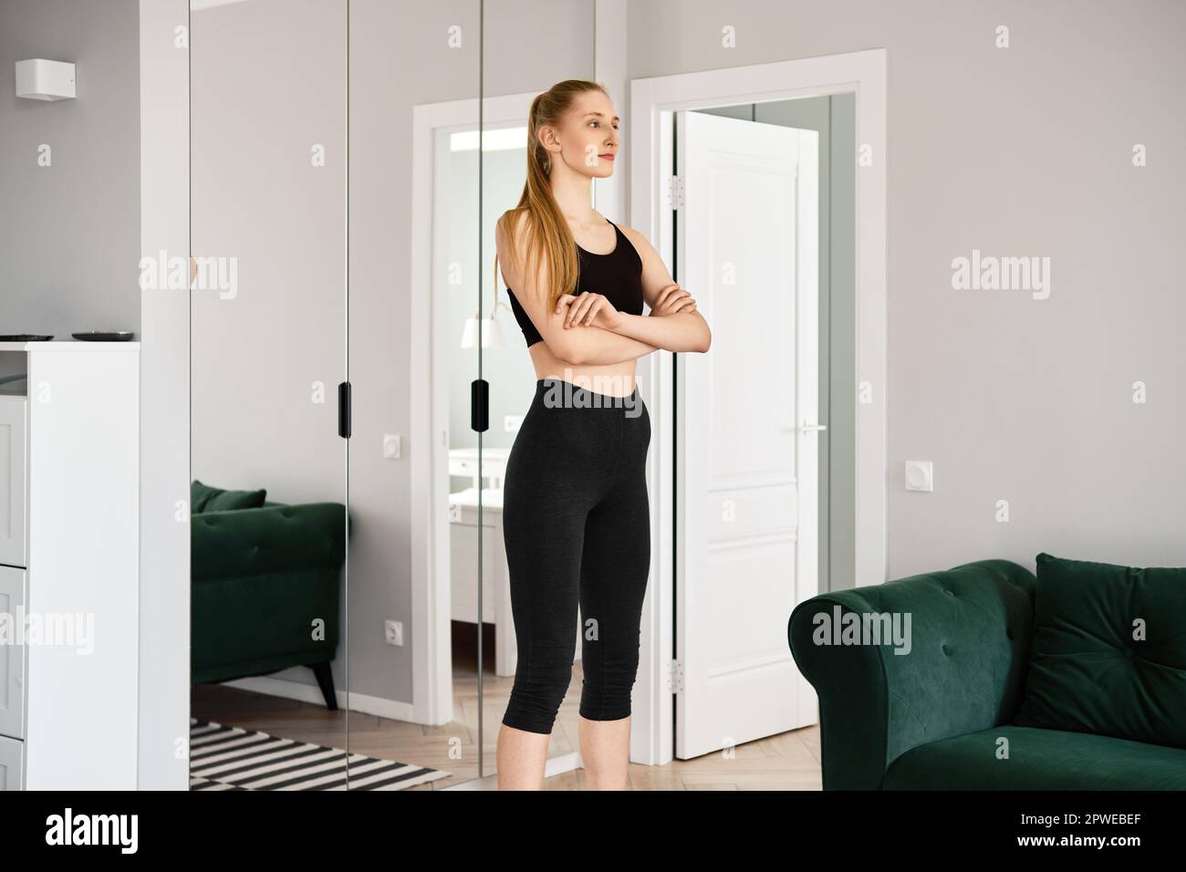 Giovane donna in abbigliamento sportivo si alza in salotto con le braccia incrociate sul petto Foto Stock