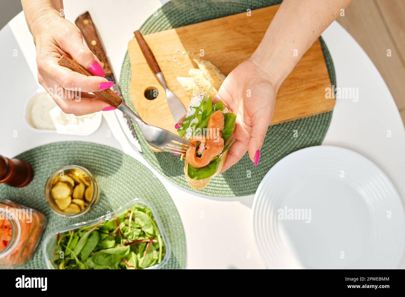 Vista dall'alto delle mani di una donna di mezza età che mette il salmone salato su un panino mentre fa colazione Foto Stock