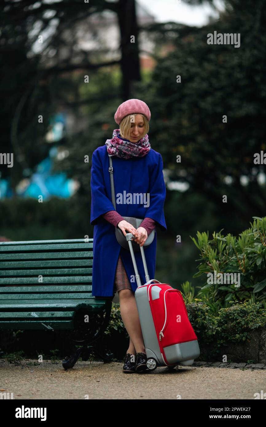 Una donna con una valigia rossa si trova vicino a una panca. Foto Stock