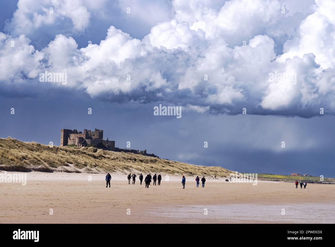Northumberland Beach; persone che camminano sulla Bamburgh Castle Beach con nuvole di tempesta sopra Bamburgh Castle, Bamburgh Northumberland Regno Unito; meteo Regno Unito. Foto Stock