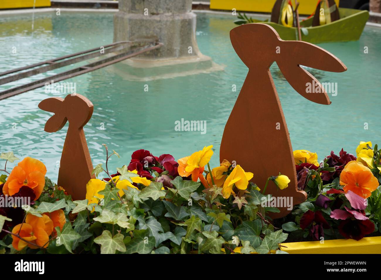 Due coniglietti pasquali all'aperto, grandi e piccoli, in legno e collocati tra i fiori. C'è acqua di fontana sullo sfondo. Foto Stock