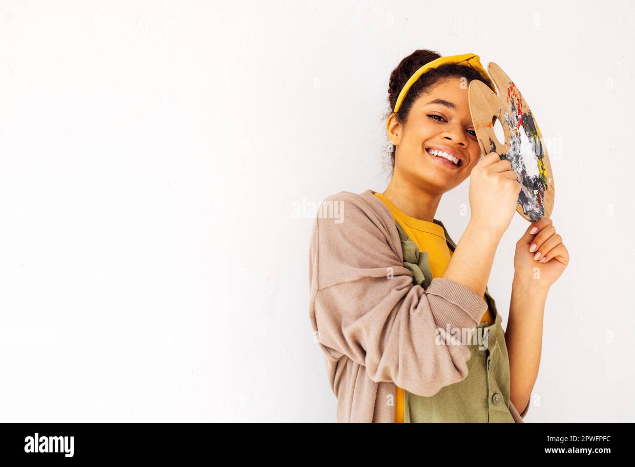 Bella donna di etnia africana sorridente posa con tavolozza di legno vicino al suo volto. Felice giovane ragazza artista ridente in abiti casual su Whi Foto Stock