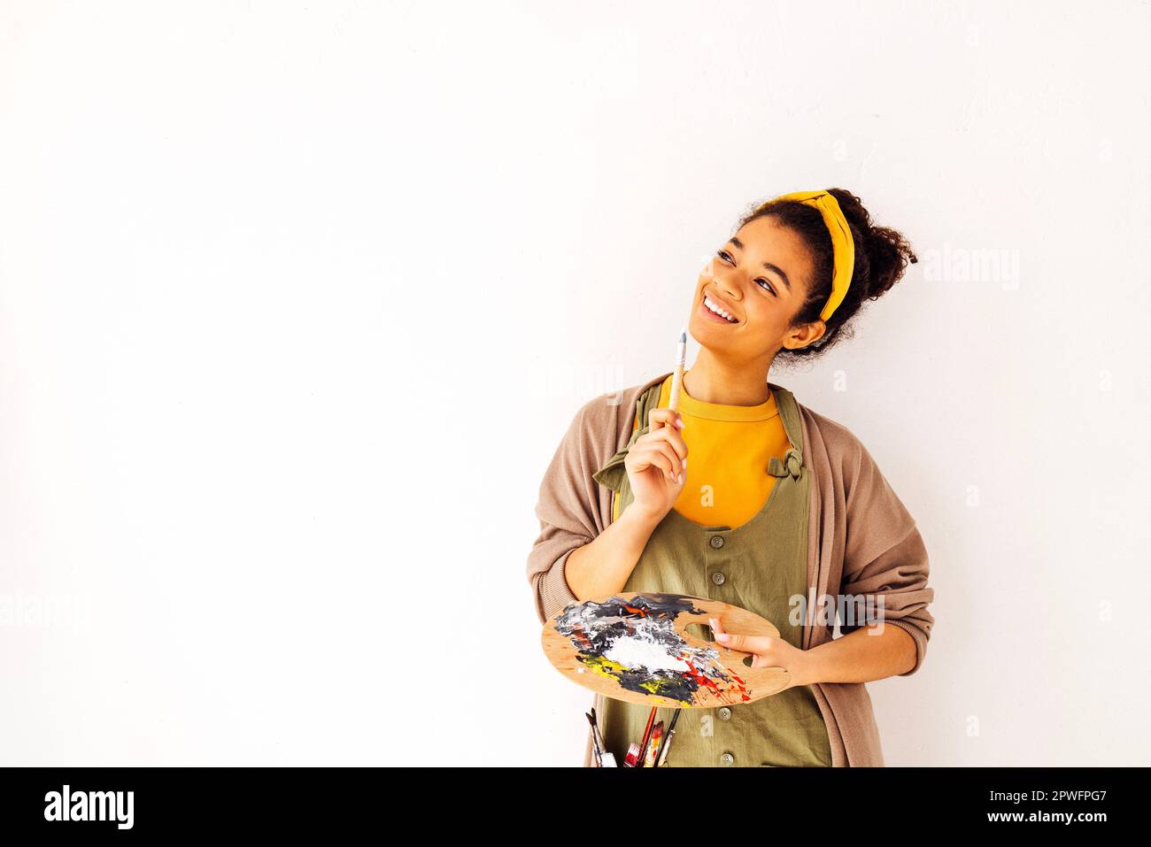 Bella donna di etnia africana sorridente posa con tavolozza di legno vicino al suo volto. Felice giovane ragazza artista ridente in abiti casual su Whi Foto Stock