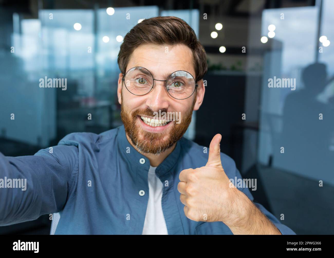 Primo piano. Un giovane uomo freelance, un blogger in occhiali, parla con la fotocamera, sorride, mostra una mano super, registra un video, blog. Foto Stock
