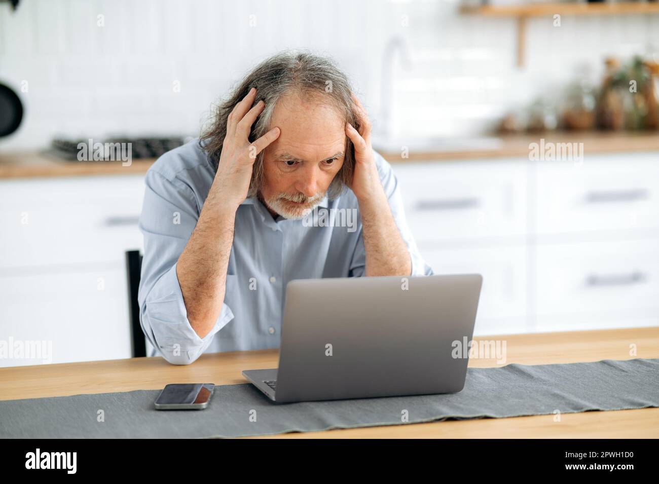 Confuso perplesso maturo grigio capelli pensionato maschio, seduto al tavolo in cucina, sorpreso, deluso guardando lo schermo del computer portatile, sconvolto dalle notizie, messaggio o risultato del lavoro Foto Stock