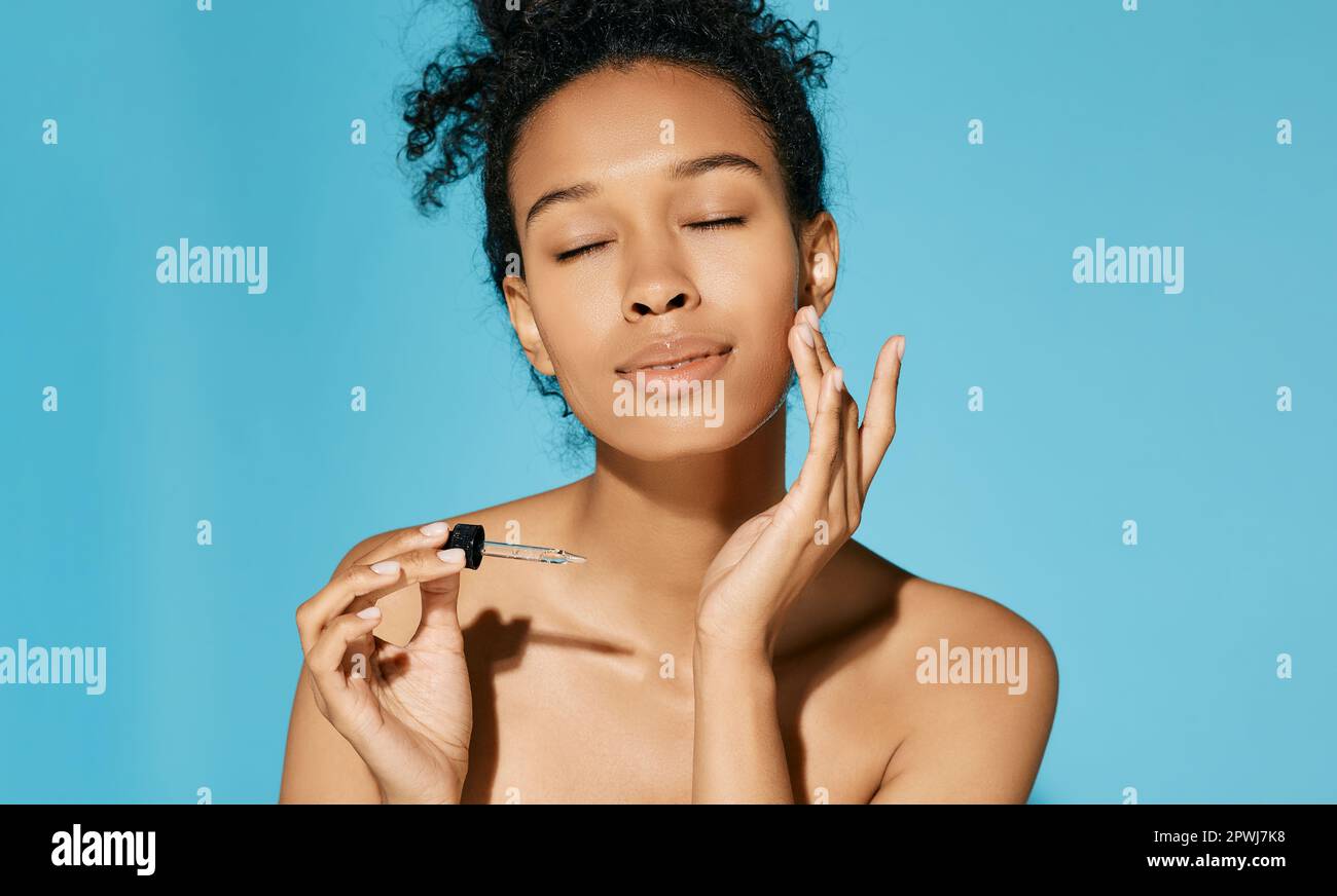 L'afroamericana le applica il siero sul viso con una pipetta, su sfondo blu. cura della pelle del viso con olio cosmetico e siero per il trattamento Foto Stock