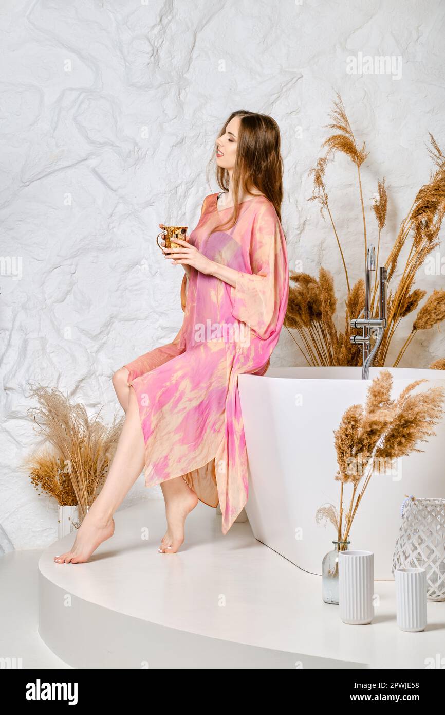 Donna carina in stola di seta con una tazza di caffè del mattino in mano si siede sul bordo della vasca da bagno. Sta andando prendere il trattamento di erbe aromatherapy della stazione termale. Foto Stock