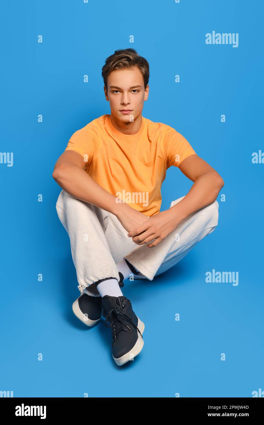 Il giovane uomo in maglietta arancione e pantaloni bianchi si siede sul pavimento dello studio su sfondo blu Foto Stock