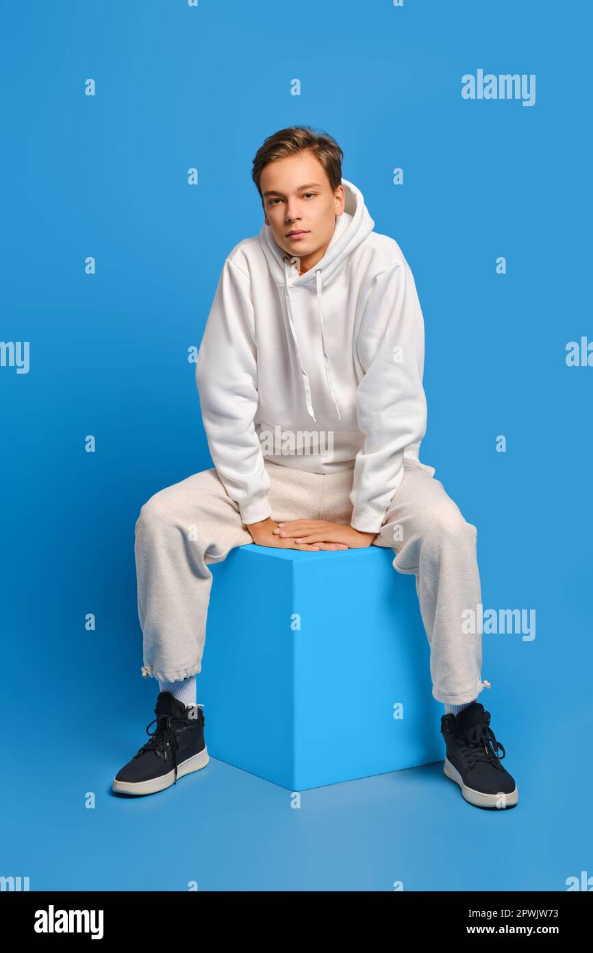 L'uomo allegro in felpa bianca e pantaloni felpa siede su un cubo di legno sullo sfondo blu dello studio Foto Stock