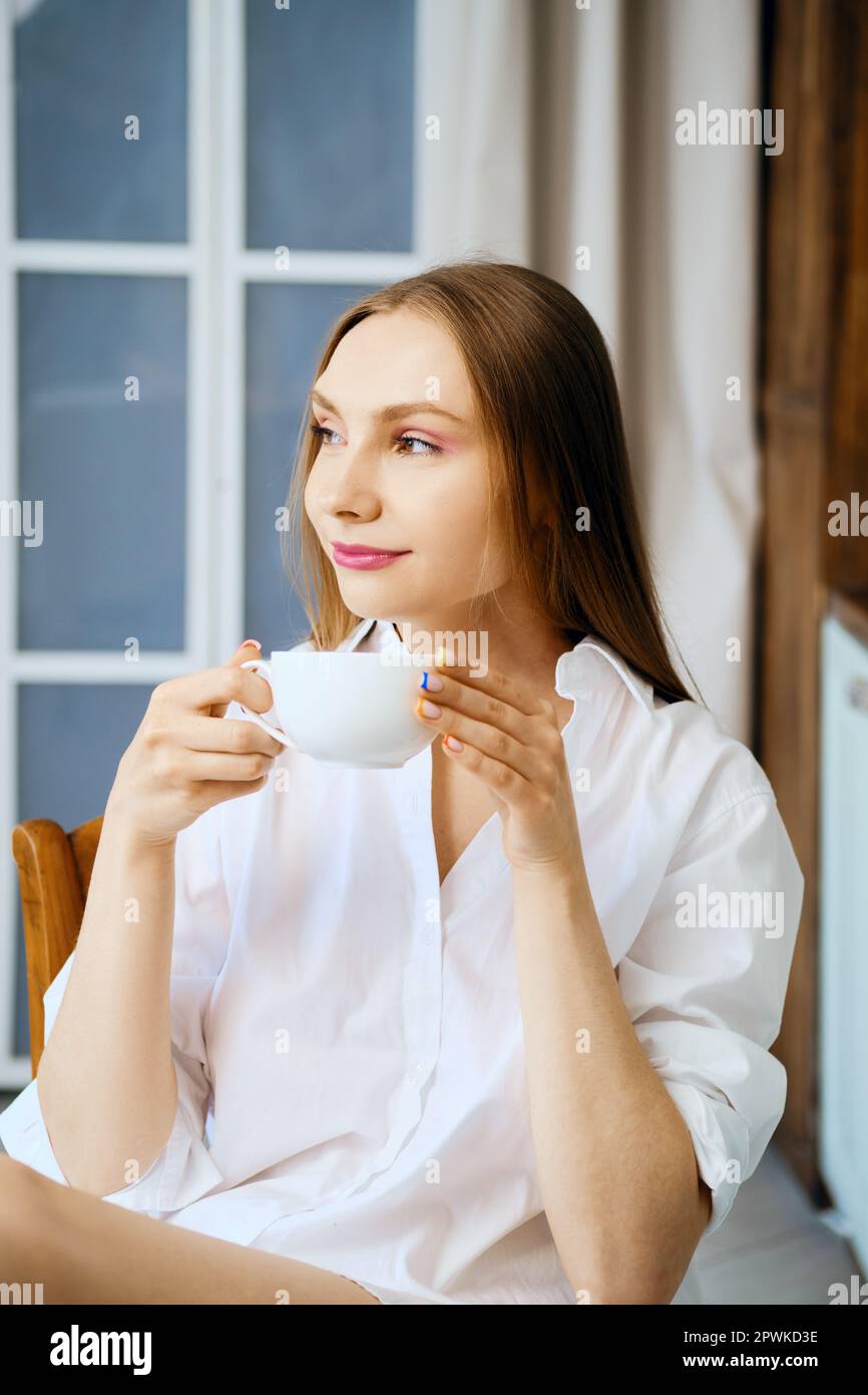 Una giovane donna in camicia bianca in cucina tiene una tazza di caffè Foto Stock