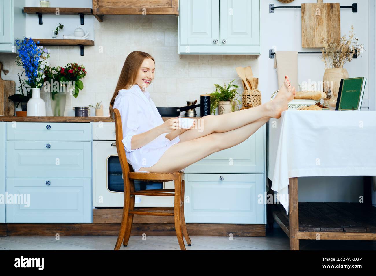 La donna che ride ha gettato le gambe a piedi nudi sul tavolo da cucina e tiene in mano una tazza di caffè Foto Stock