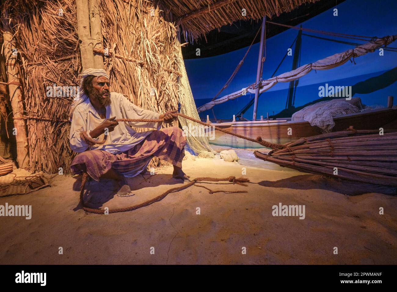 Un pescatore che tira una linea di corda dalla sua rete, dispositivo di cattura di legno sulla spiaggia, barca a vela sullo sfondo. Un diorama raffigurante una scena del passato degli Emirati Arabi Uniti, Foto Stock