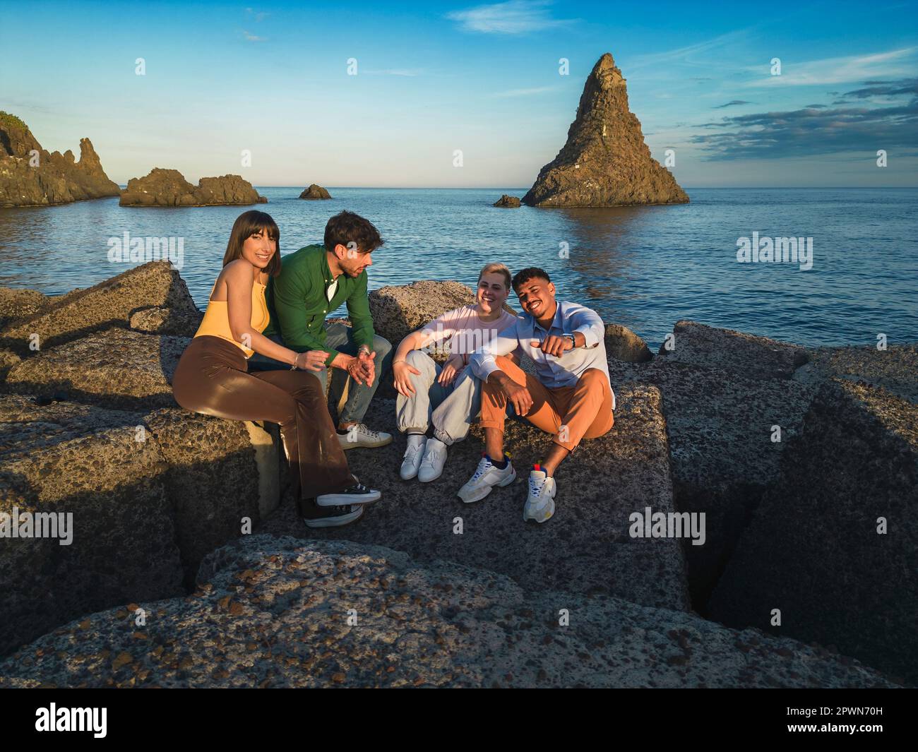 Quattro diversi amici seduti sui blocchi del porto di Acitrezza, il drone aereo ha girato. Isola di Lachea e una pila sullo sfondo. Foto Stock