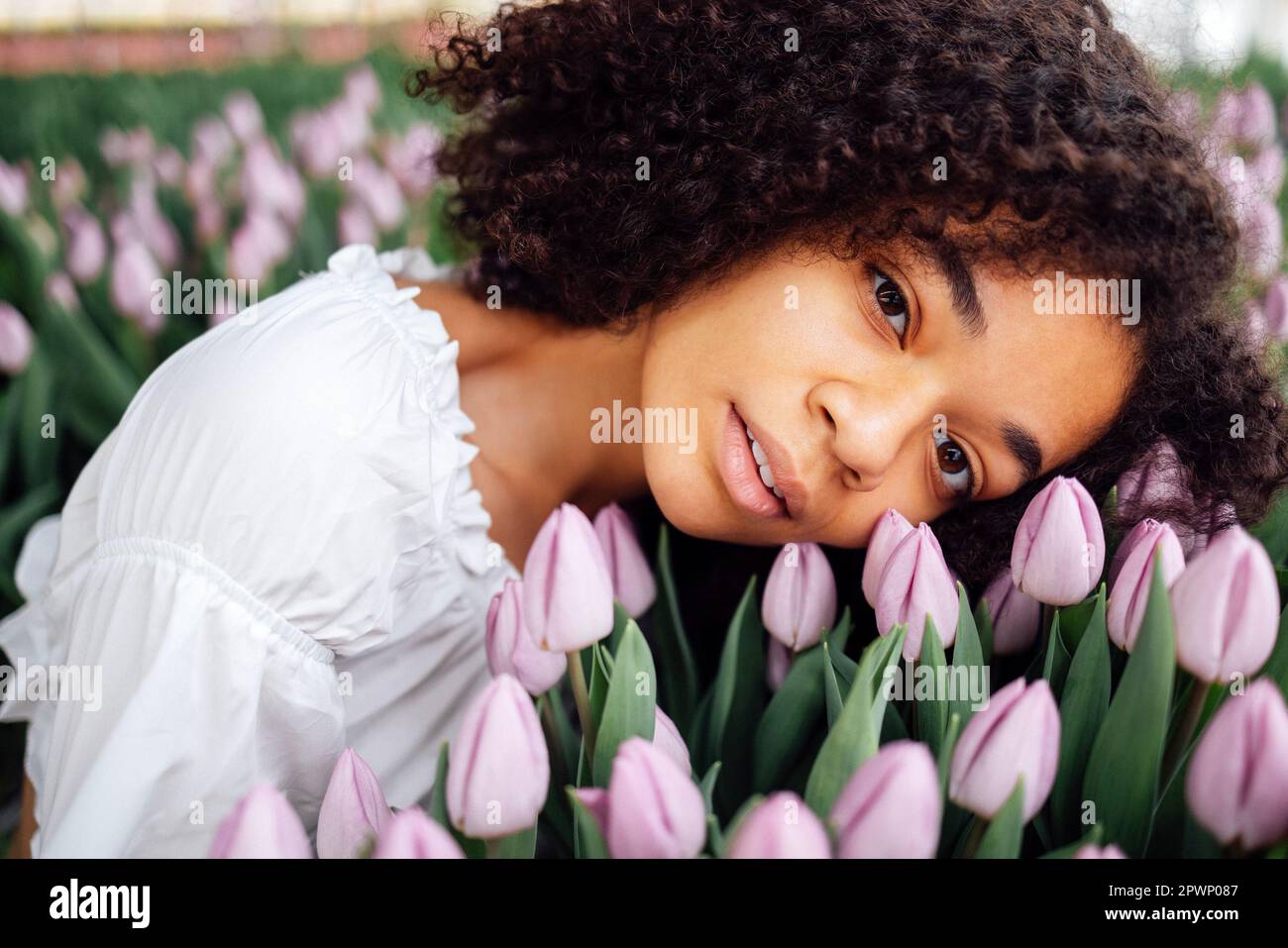 Primo piano di ritratto femminile di carina affascinante ragazza etnica africana tra boccioli di tulipano di colore delicatamente viola. Giovane ragazza afro romantica in maglietta bianca touc Foto Stock