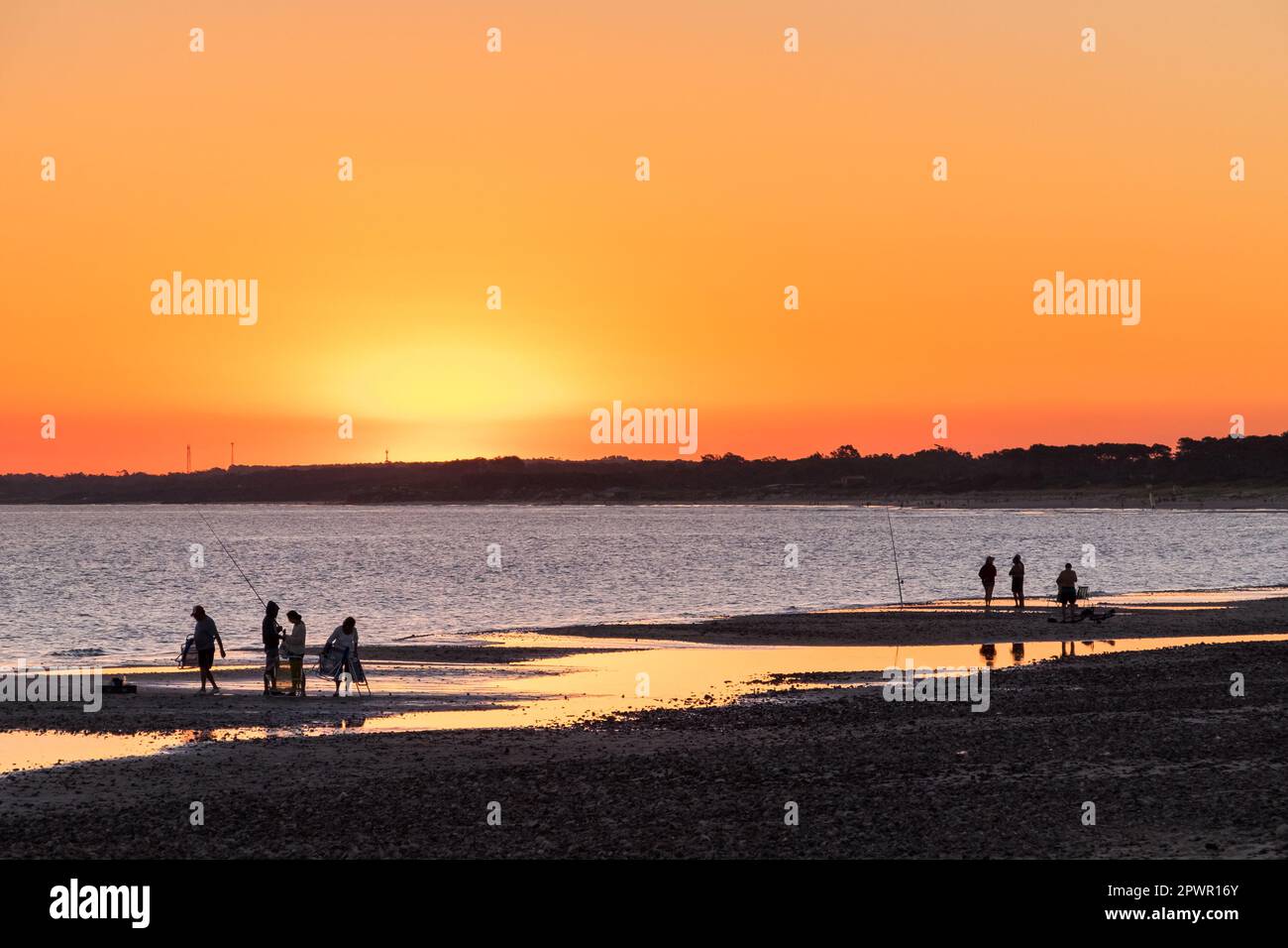 Sagome di pescatori sulla spiaggia di Las Flores, durante un tramonto rossastro, con alcune pozze d'acqua sulla sabbia Foto Stock