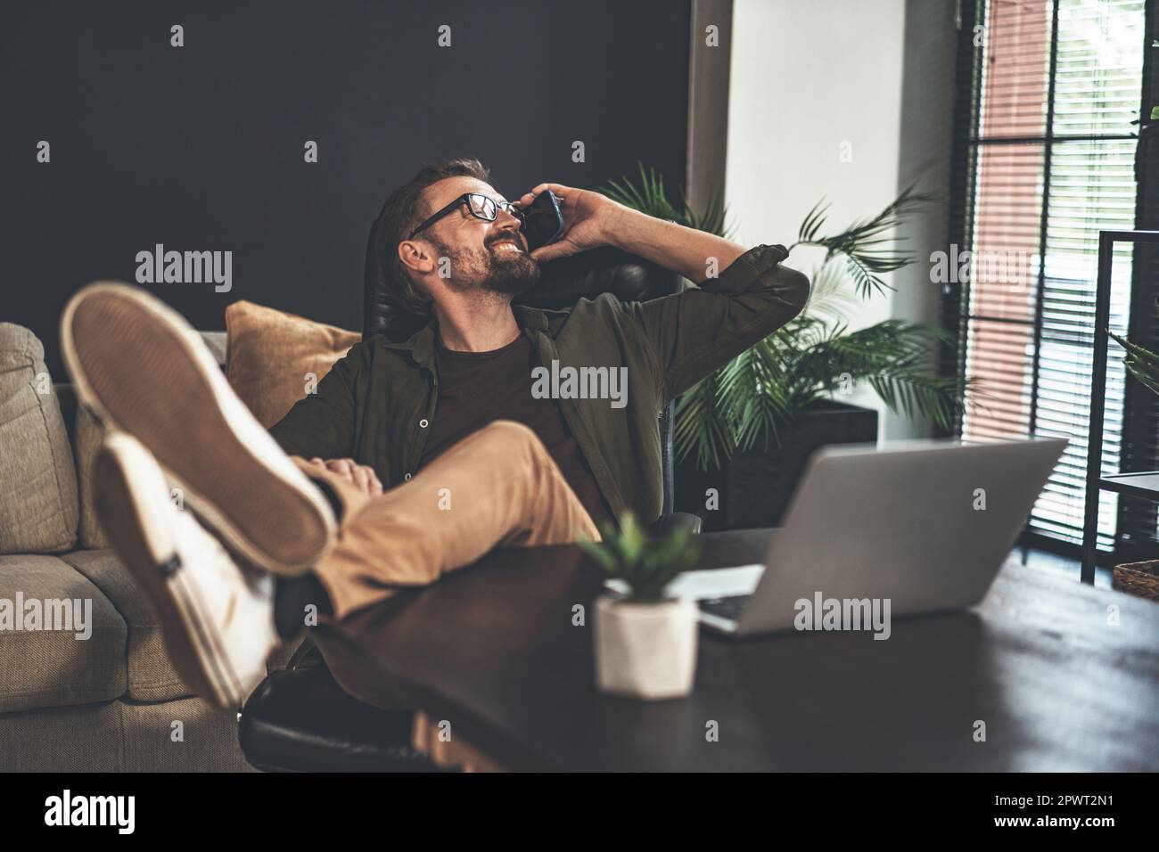 Un freelance felice e rilassato lavora da casa e prende una telefonata mentre si siede alla scrivania con i piedi in alto come un cowboy. Guarda fuori dalla finestra con un sorriso sul viso, godendo della flessibilità del lavoro remoto e del comfort della propria casa. Foto di alta qualità Foto Stock