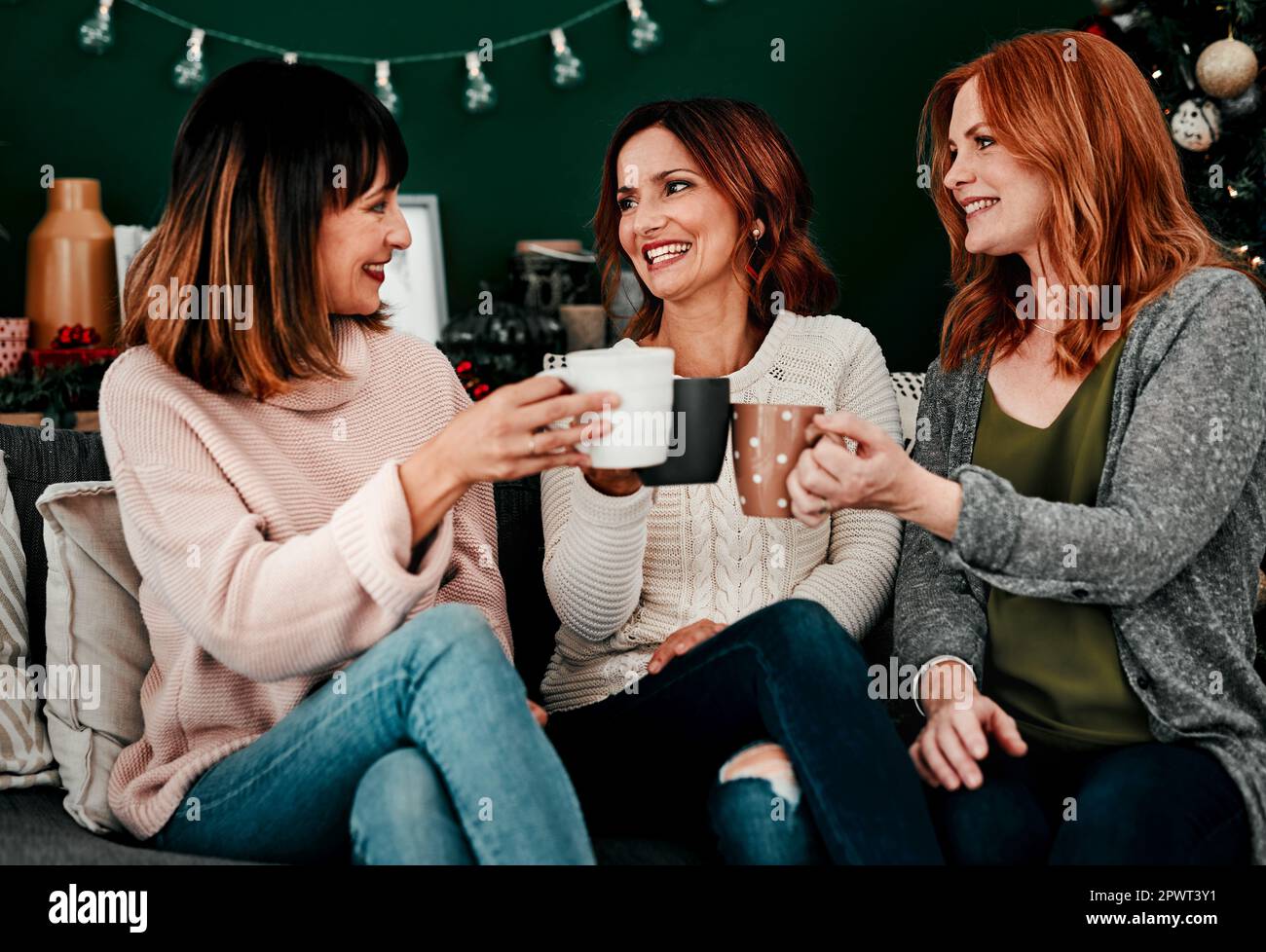 Heres a noi Signore. Tre donne di mezza età sedette insieme su un divano mentre bevono il caffè a casa durante il periodo natalizio Foto Stock