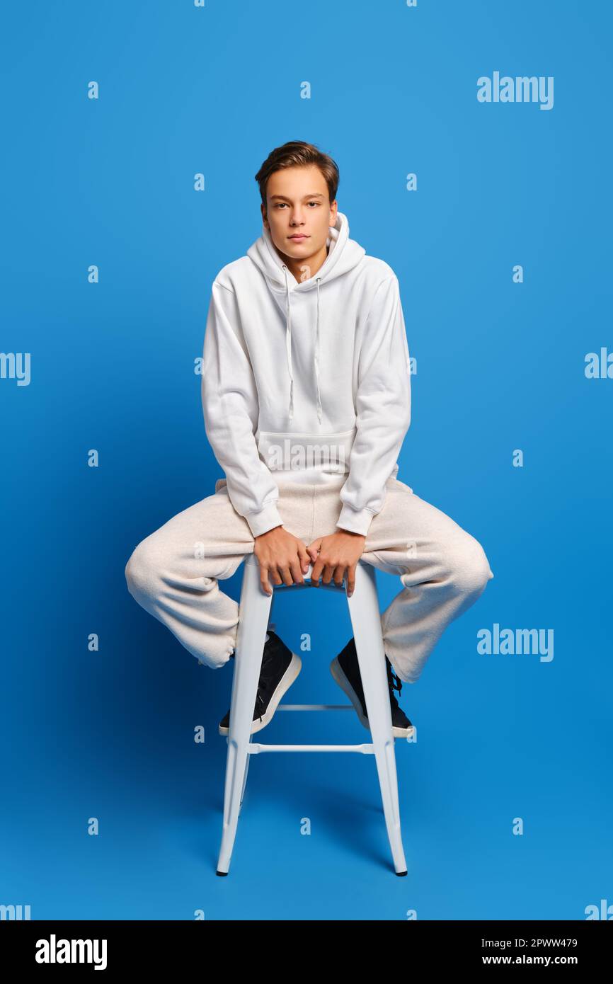 Bell'uomo che indossa una felpa bianca e pantaloni sudoripiti seduti su una sedia bianca sullo sfondo blu dello studio Foto Stock