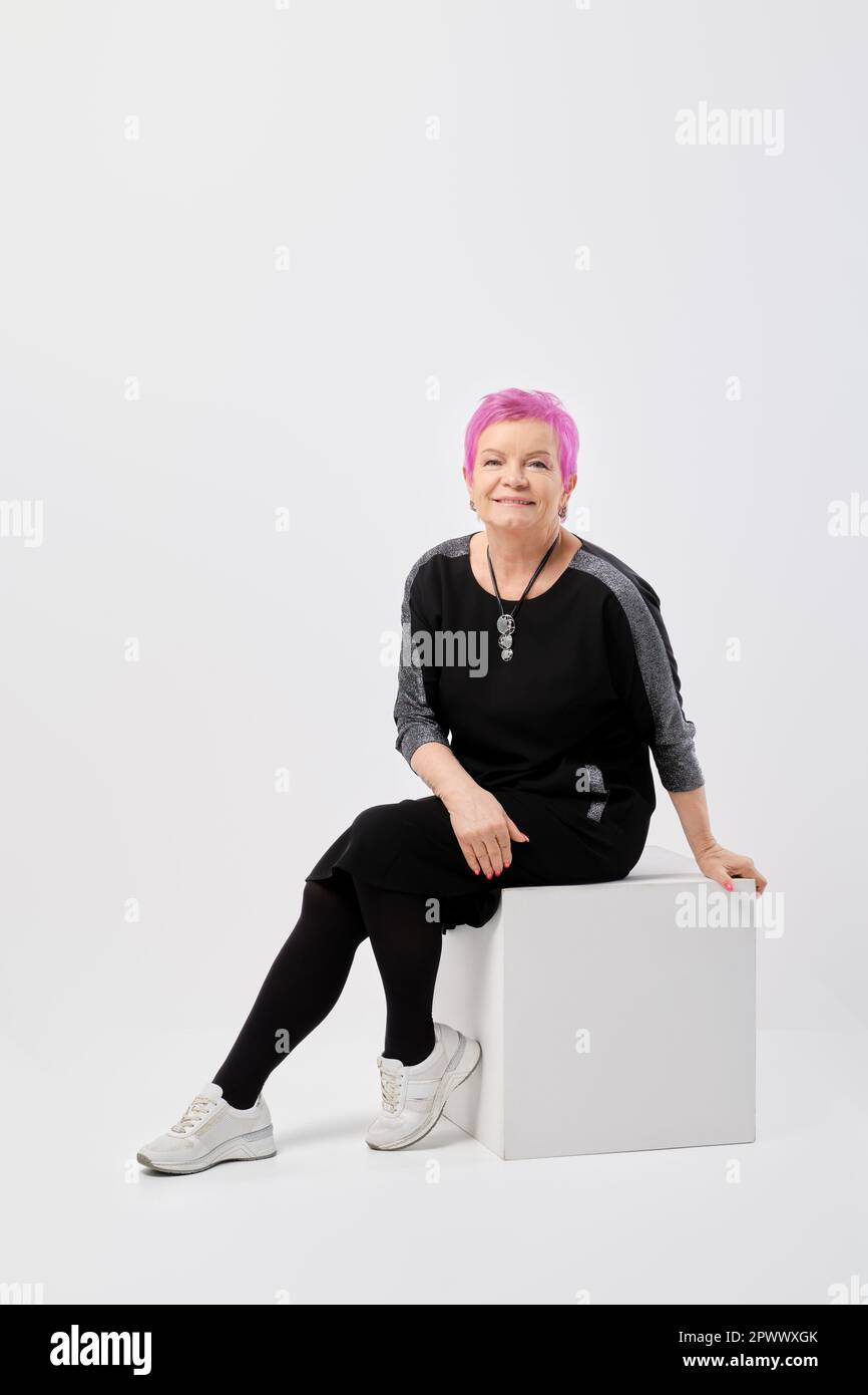Felice vecchia donna con i capelli corti rosa nel vestito a maglia lungo siede sulla scatola bianca che si appoggia in avanti nello studio Foto Stock