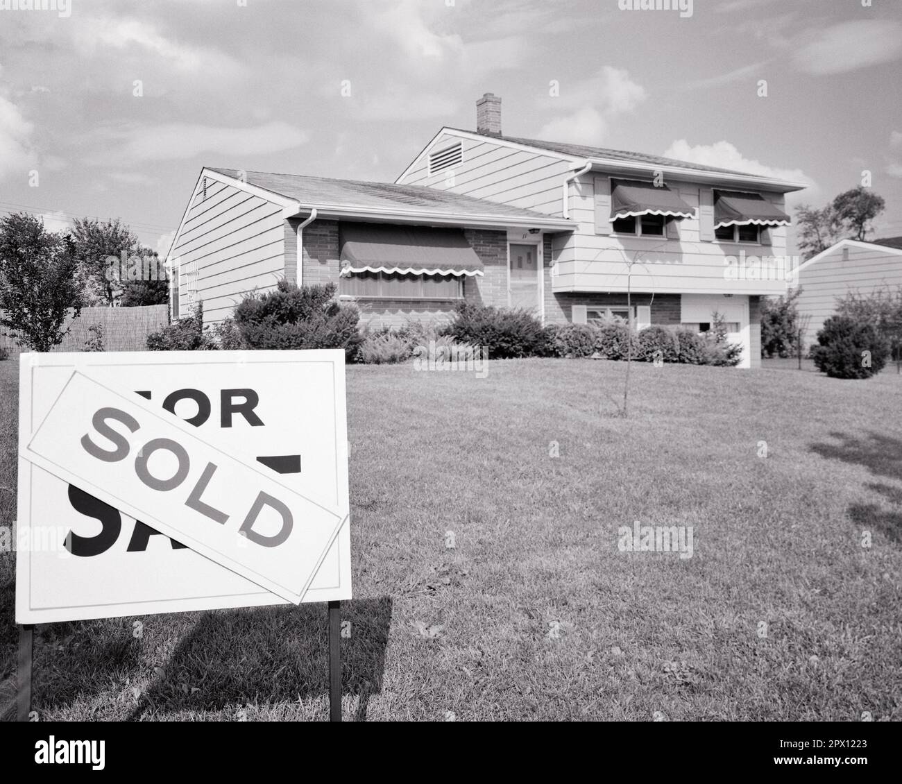 1960S CASA SUBURBANA A LIVELLO DIVISO CON SEGNO VENDUTO NEL CORTILE ANTERIORE - B22878 HAR001 HARS CONCETTI SIMBOLICI A LIVELLO DIVISO LIVELLO BIANCO E NERO HAR001 RAPPRESENTAZIONE VECCHIO STILE Foto Stock