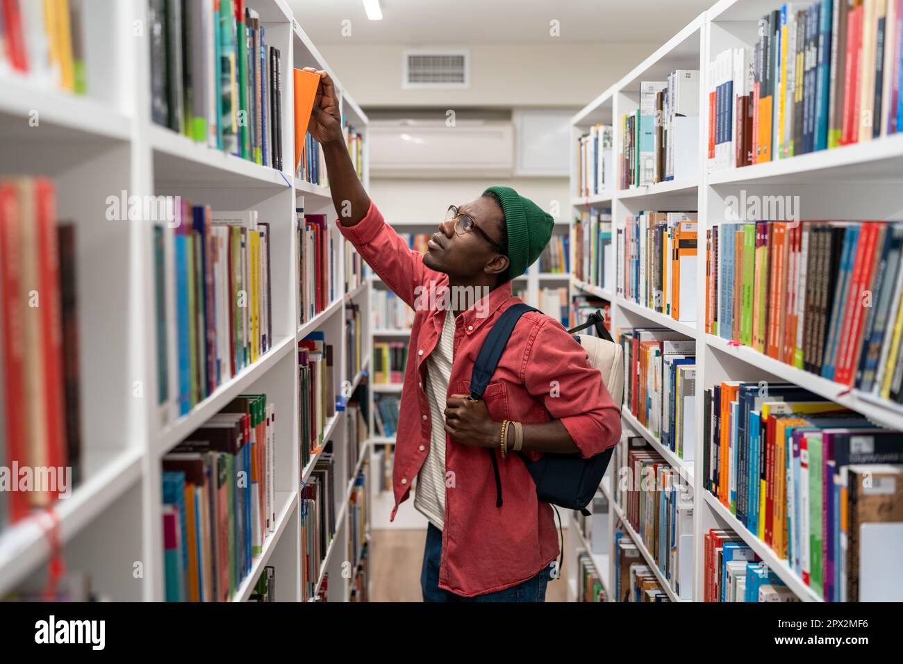 Allievo nerd uomo afroamericano che sceglie il libro nella biblioteca universitaria prendendolo dallo scaffale. Foto Stock