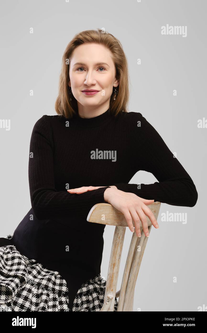 Donna sorridente in dolcevita seduta su sedia alta appoggiata alla schiena in studio Foto Stock