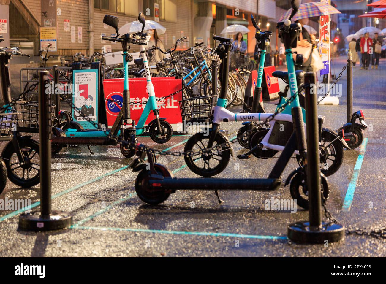 Osaka, Giappone - 29 aprile 2023: Forte pioggia su mini bici elettriche condivise e scooter parcheggiati sulla strada cittadina Foto Stock