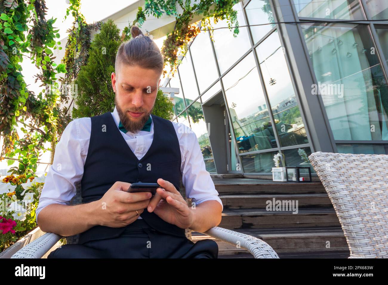 Giovane hipster in una camicia bianca, gilet nero guarda in uno smartphone mentre si siede in un attico Foto Stock