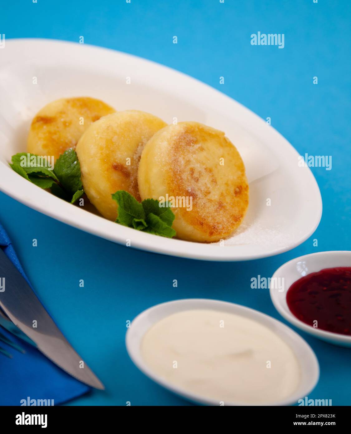 Frittelle di formaggio ricotta rotonda in un piatto bianco su sfondo blu. Cibo prima colazione. Cibo sano. Salsa di panna acida e menta. Primo piano vista dall'alto. Pasto dessert. Frittelle di ricotta al formaggio ricotta. Foto Stock