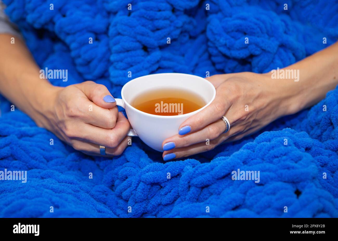 Una ragazza avvolta in una grande coperta maglia a mano di lana merino blu,  una coperta calda e molto morbida, una ragazza che tiene una tazza bianca  di tè caldo, posto per