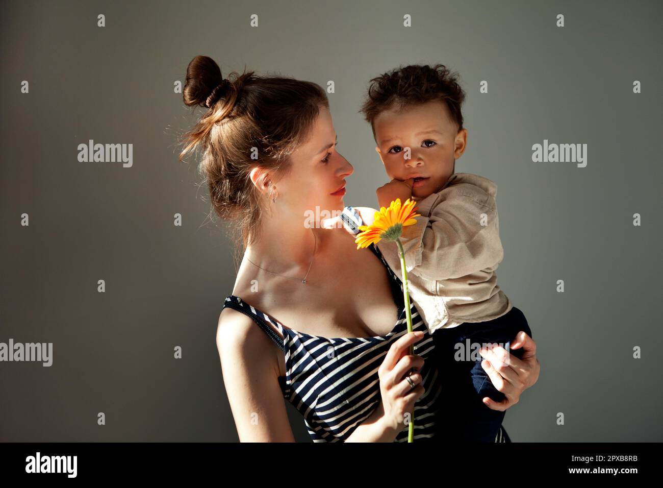 Ritratto bella Madre tiene in mano il bambino e il fiore gerbera mentre il bambino guarda la macchina fotografica. Autentico ritratto di vera famiglia, soleggiato, duro ligh Foto Stock